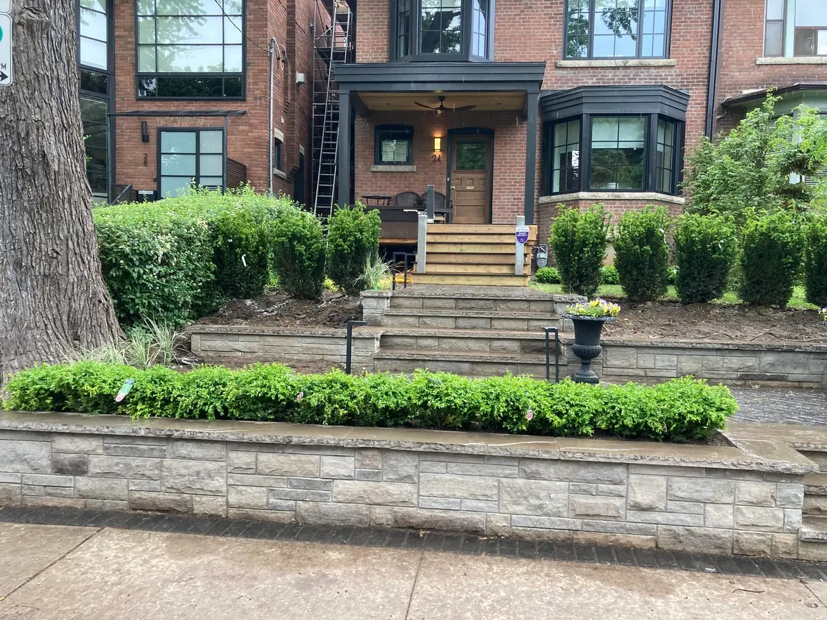 A retaining wall and steps to a brick house porch, with trimmed hedges.