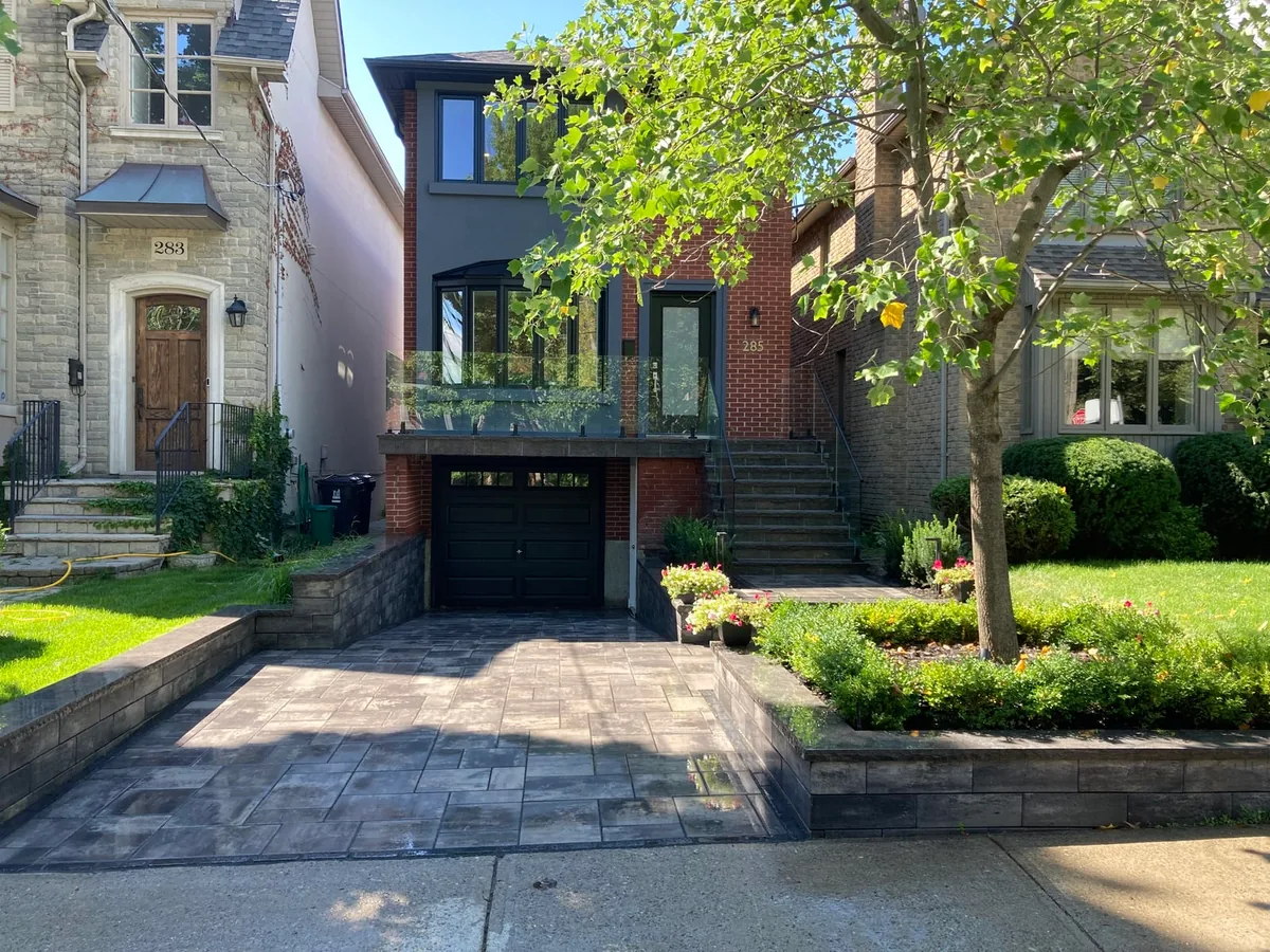 A stone-clad house front with a black garage door, a paver driveway, retaining-wall planters, and a glass-railed balcony above.