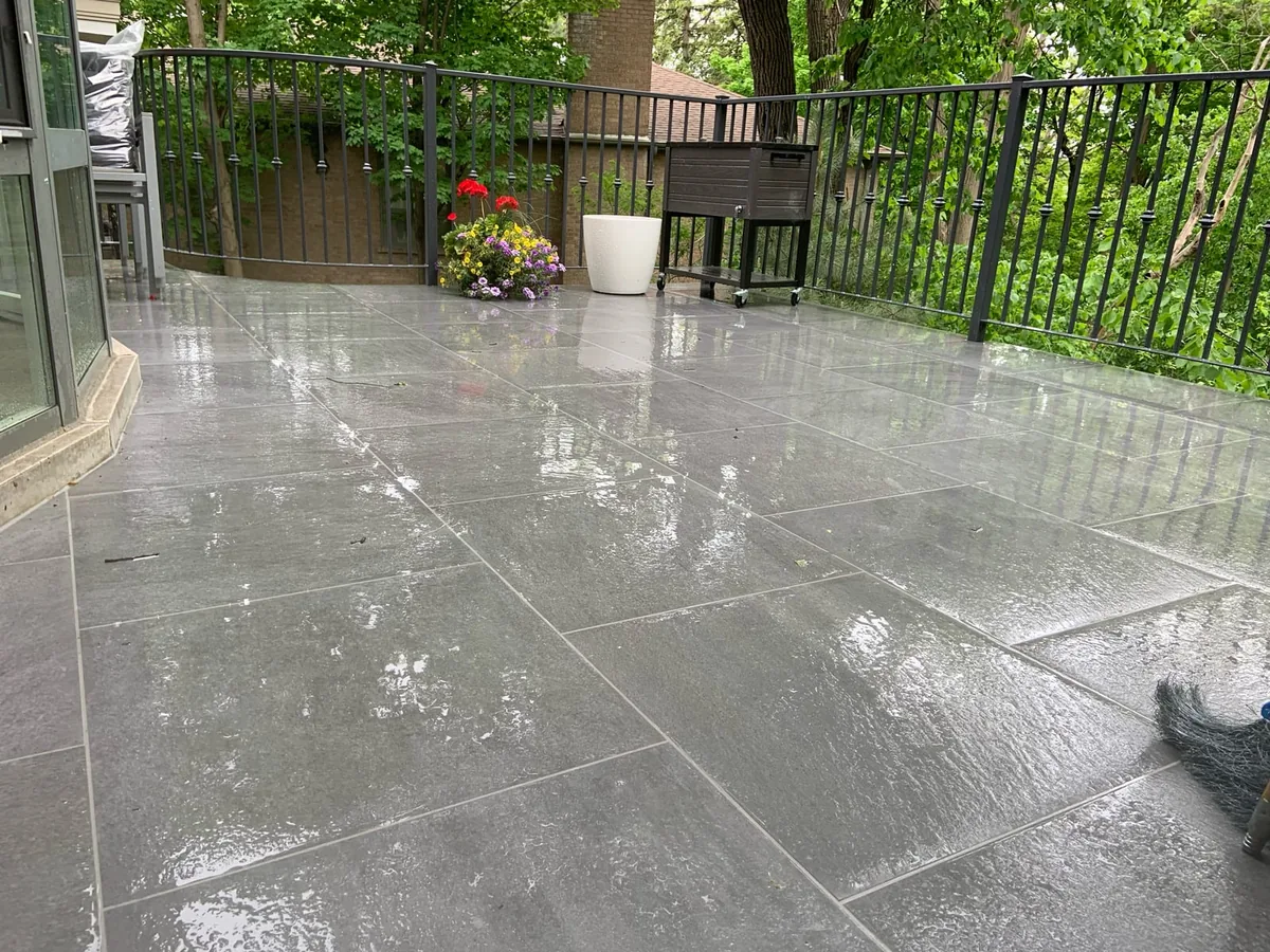 A rainy balcony patio with a grill covered in plastic and a wire hanging basket of flowers.