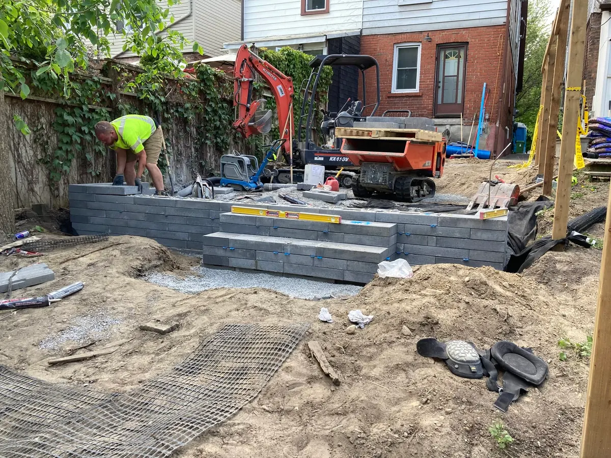 A backyard retaining wall being built with large segmental concrete blocks, a mini excavator, and a wood fence covered in ivy.