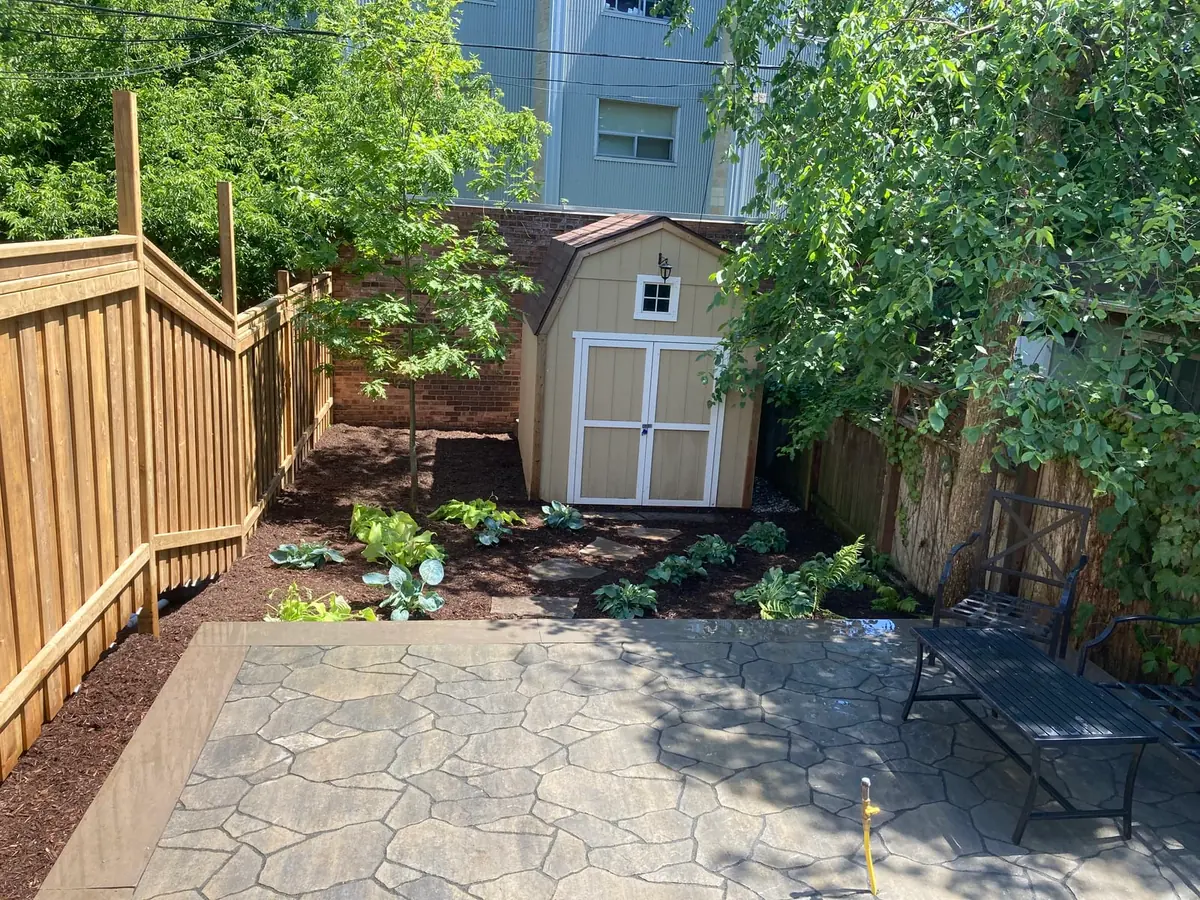 A backyard garden bed with hostas and ferns beside a wood fence and shed, with an iron bench.