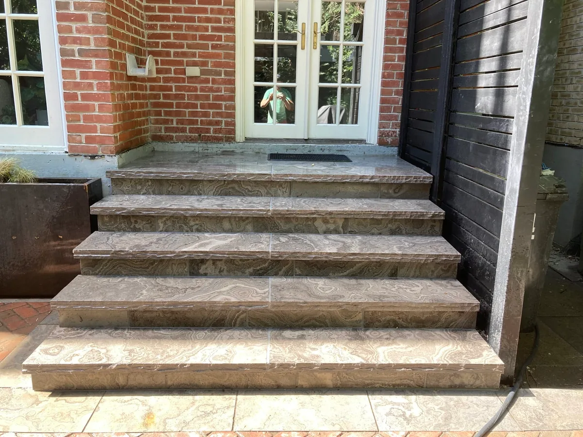 Marble-look steps at a brick house beside a dark slat fence and a garbage bin.