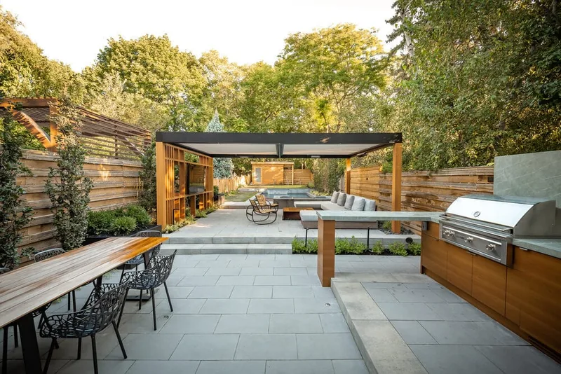 A backyard patio with large pavers, a wood pergola with slatted screens, built-in bench seating, and an outdoor kitchen counter with a grill.