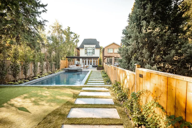 A backyard rectangular pool with stone coping, a pergola structure, and a stepping-paver path across the lawn.