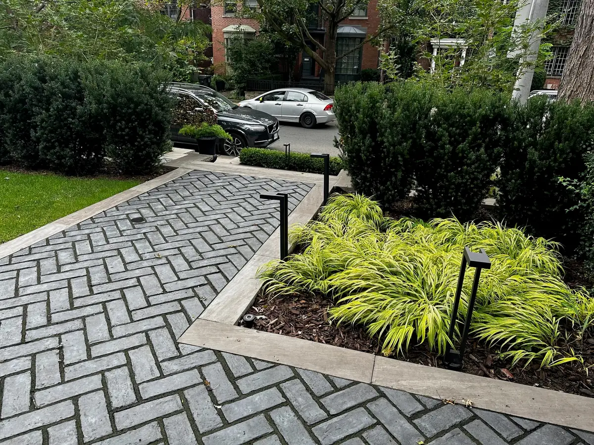 A herringbone-pattern paved driveway with a car parked beside shrubs.