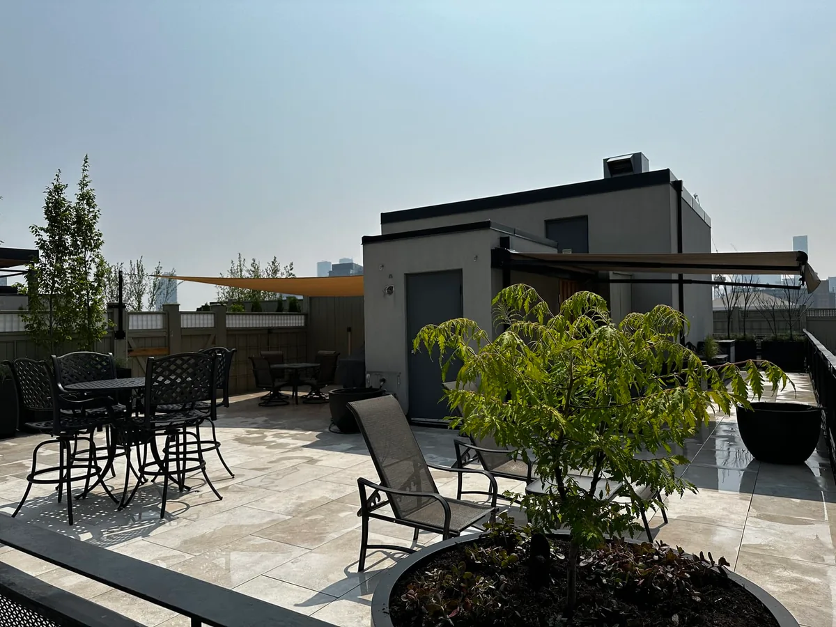 A rooftop terrace with a bar-height table and chairs, a small storage building with an extended awning, and a hazy skyline in the distance.