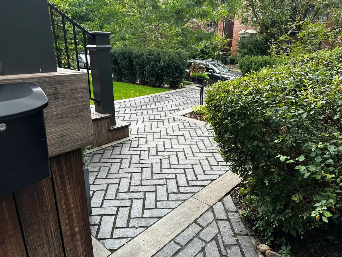 A paved walkway near a porch with a mailbox post and a hedge.