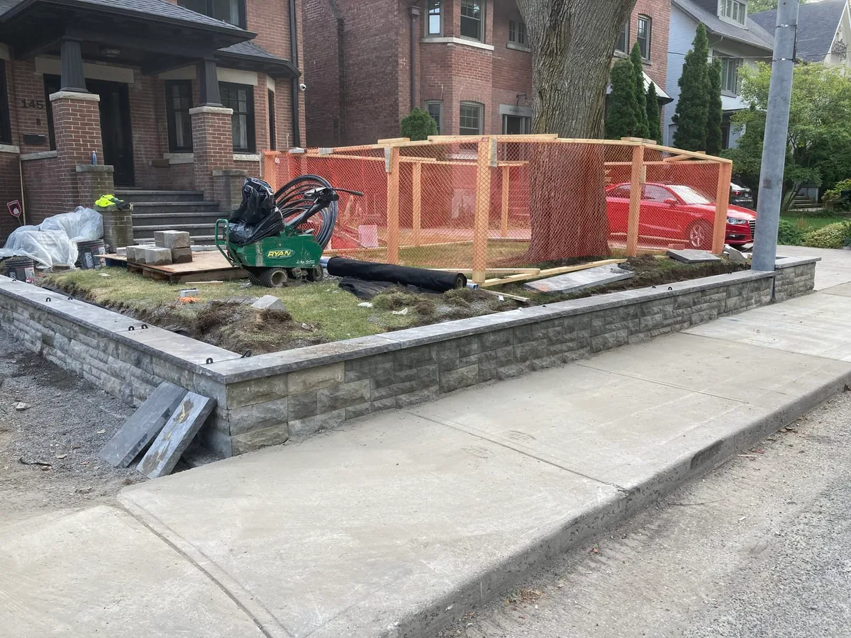 A front yard with a new stone-look block retaining wall around a lawn bed, a small trenching machine, and orange safety fencing.