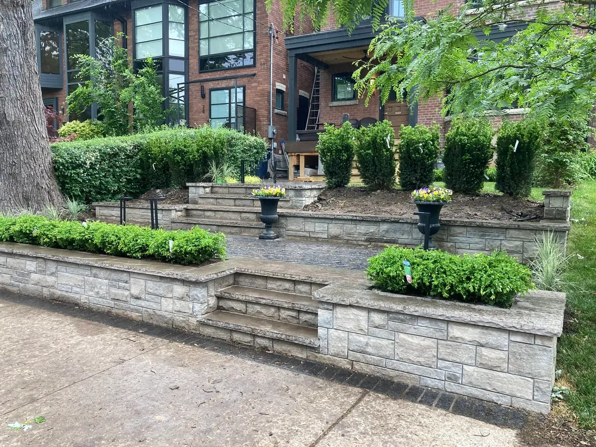 A front-yard retaining wall with hedges, urns of flowers, and steps leading to a house porch.
