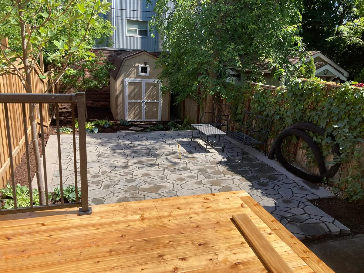 A wood deck foreground looking onto a flagstone patio with an iron bench and an ivy-covered fence trellis.