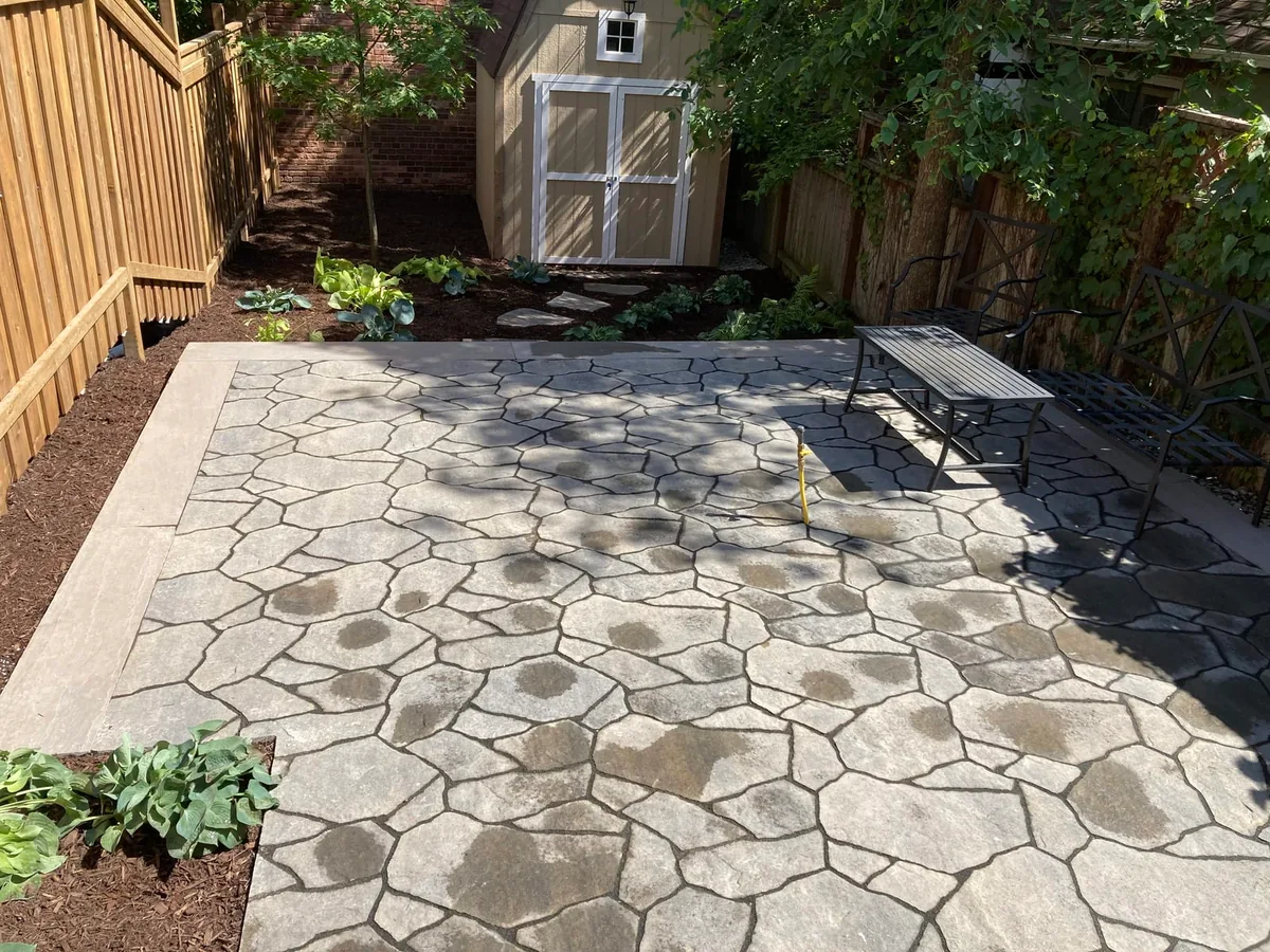 A wider view of the flagstone patio, wood fence, shed, and iron bench in dappled light.