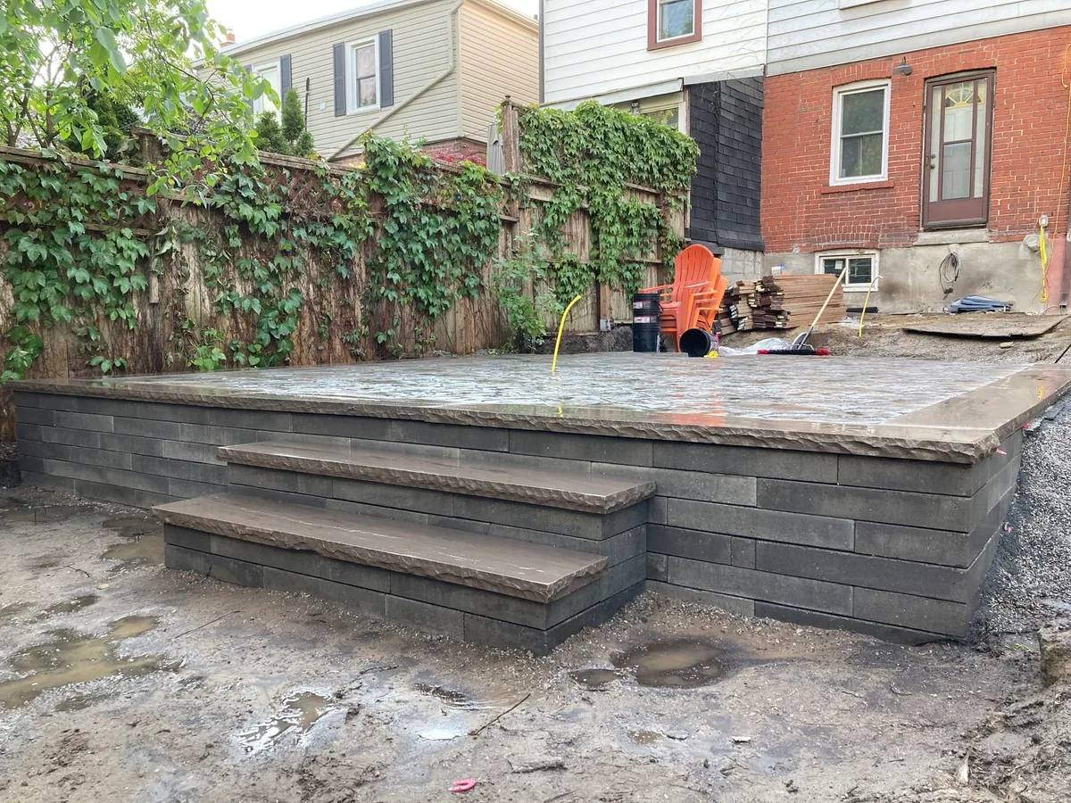 A stone-clad retaining wall with built-in steps leading up to a patio, an orange chair, and a rain-wet surface.