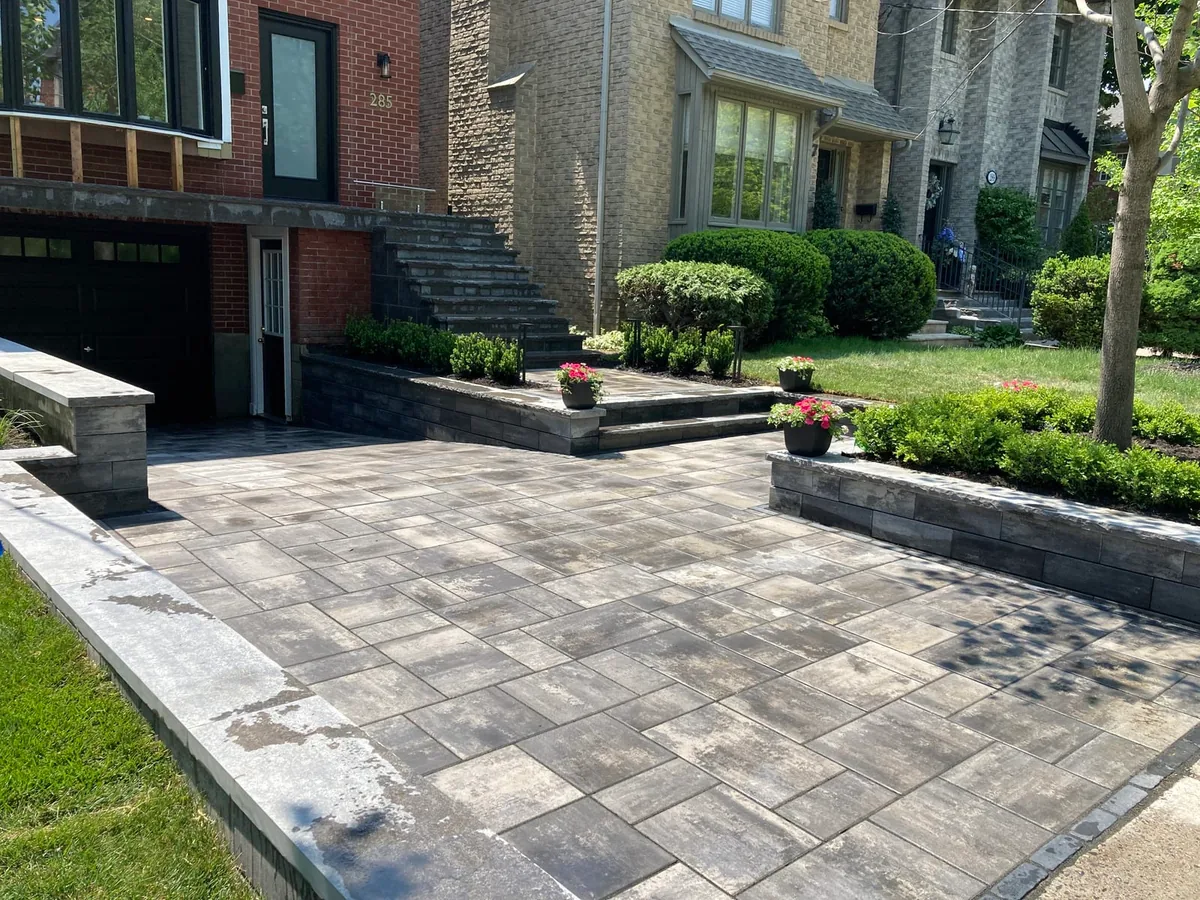 A wide view of a paver driveway and black stone steps leading to a house, with neighbouring houses visible.