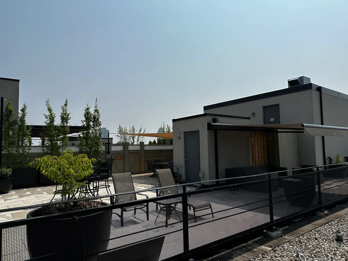 A rooftop patio with a pergola, small trees in planters, a mesh privacy railing, and a triangular shade sail.